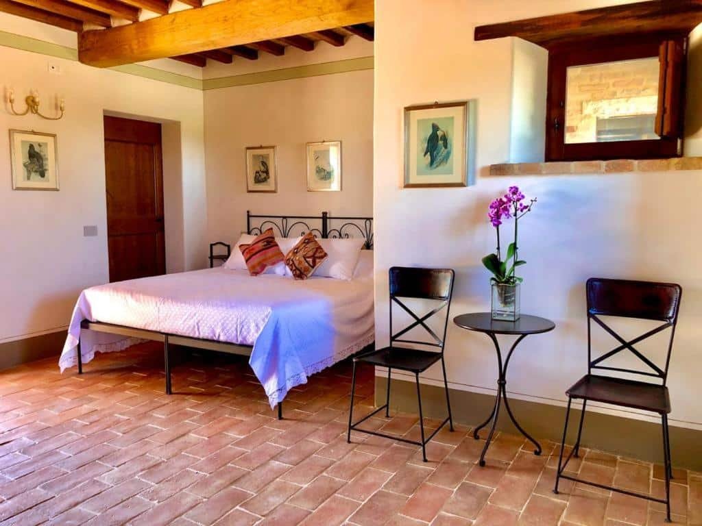 Quarto do Agriturismo Sole, com 23 m², chão de taco, com mesinha pequena redonda preta e uma cadeira preta de cada lado, um quadro em cima com desejos no tom azul, cama de casal ao lado com alguns quadros na parede. 