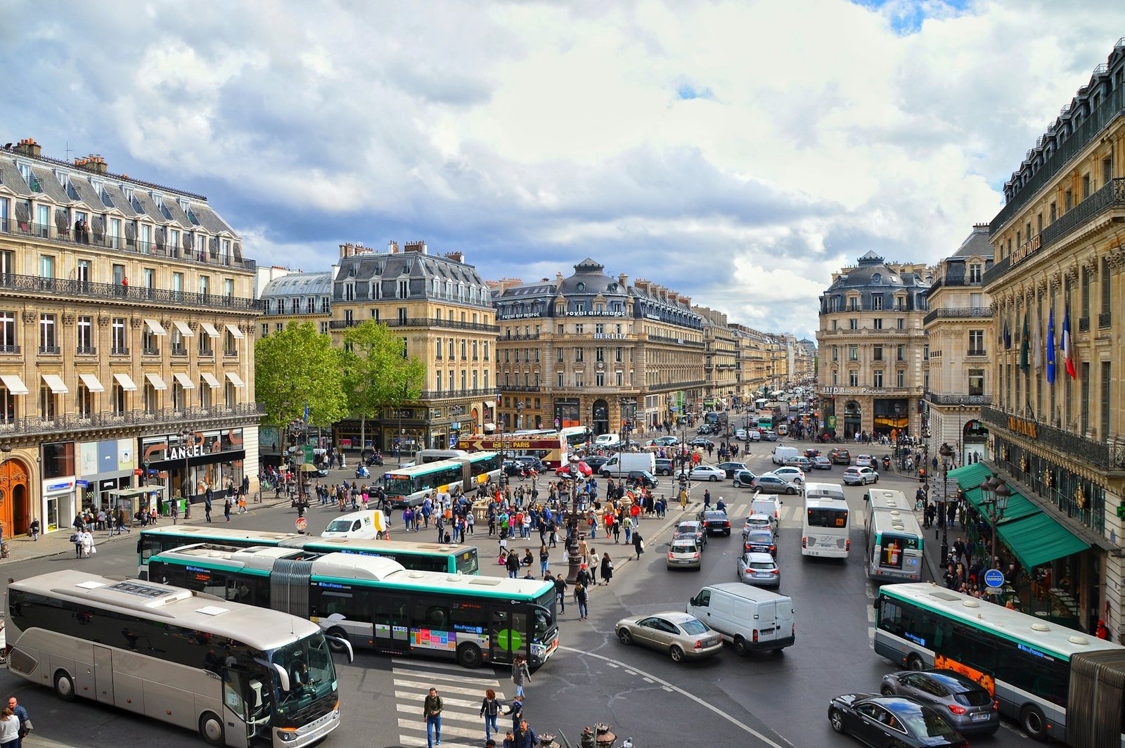 Um movimentado cruzamento da cidade com ônibus, carros e pedestres cercados por edifícios históricos sob um céu parcialmente nublado. A imagem ilustra o post sobre transportes em Paris.