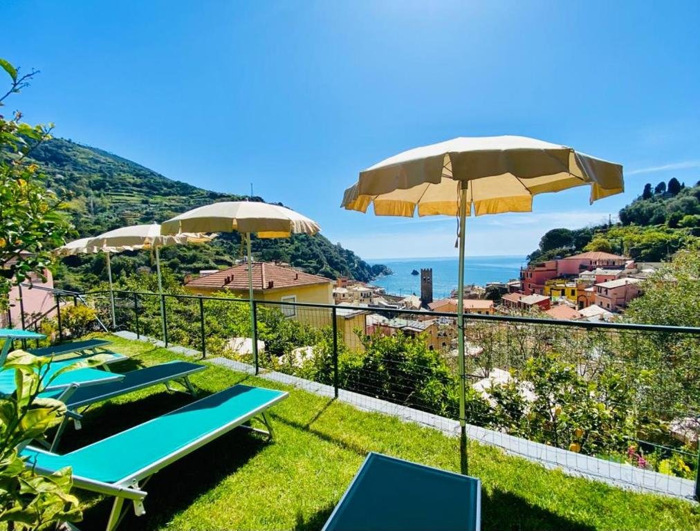 Terraço com várias cadeiras azuis em cima de um gramado verde, alguns guarda-sóis amarelos, árvores em volta e uma vista para as casas coloridas, montanhas e mar durante o dia, ilustrando post Hotéis em Cinque Terre.