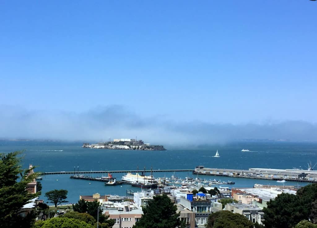 Ilha de Alcatraz do ponto de vista de San Francisco. A foto foi tirada de longe, então a ilha fica pequena no meio do mar. Em primeiro plano estão os barcos e construções de San Francisco.