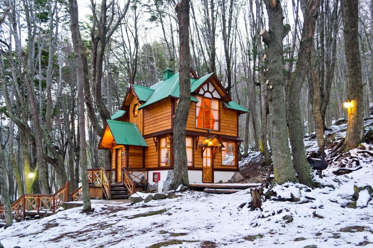 Um chalé de madeira com telhado verde é cercada por árvores nevadas em uma área florestal. Luzes quentes estão acesas do lado de fora na Aldea Nevada.