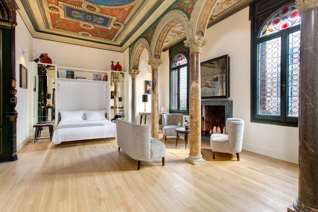 quarto do Roof Suite Rome um aluguel de temporada em Roma, em um ambiente amplo, poltronas brancas ao redor de uma mesa e cama enorme com mesinha e luminária de ambos os lados, há arcos trabalhados com afrescos que sobrem até o teto, as janelas são garndes