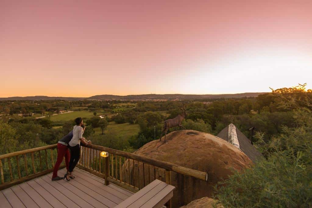 Um casal está em um deck de madeira, com vista para uma vasta savana ao pôr do sol, com uma estátua de um antílope em primeiro plano para representar um dos melhores lugares para lua de mel