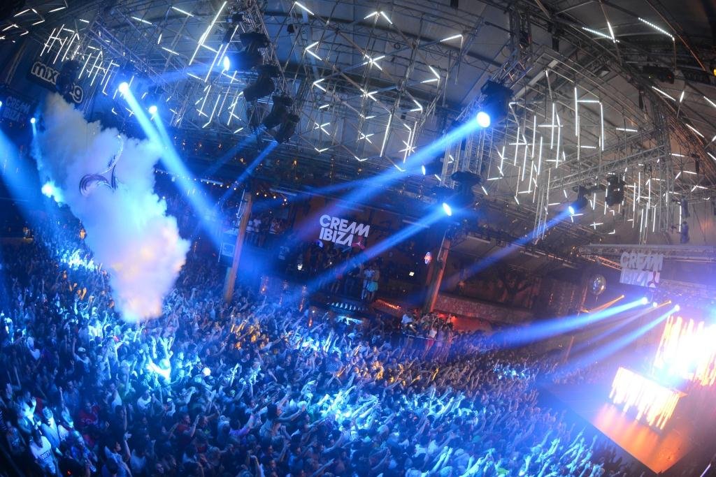 Uma grande multidão dança em uma boate vibrante com luzes brilhantes, efeitos de fumaça e "Cream Ibiza" exibido em uma tela.