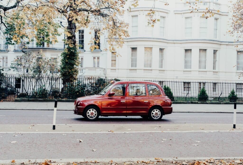 Um carro vermelho em estilo europeu passando por uma rua de Londres pouco movimentada e algumas árvores