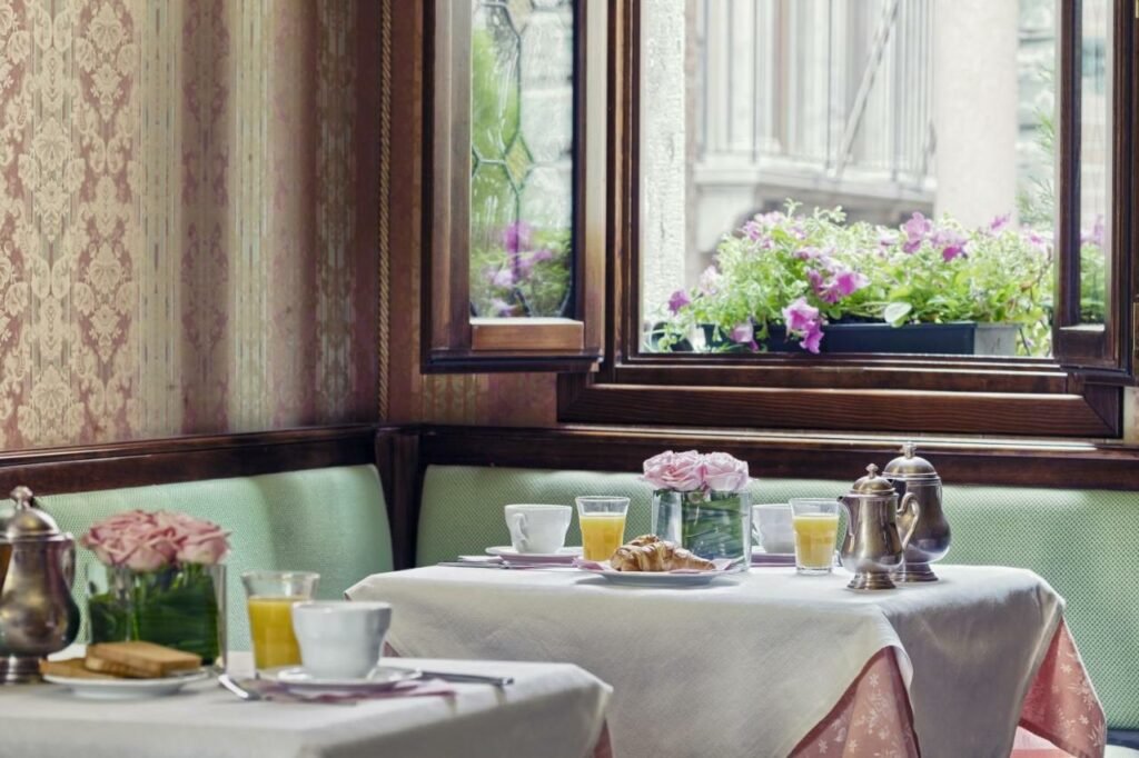 Mesas do restaurante Antica Locanda al Gambero com vista para a cidade com um vaos de flores roxas no beiral da janela, as mesas estão com toalhas vermelhas e brancas