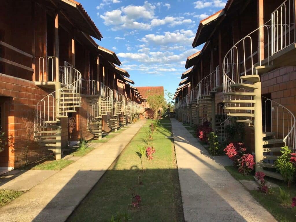 Vista de fora de todos os Apartamentos Maranduba Ville II.  Eles estão alinhados e todos de frente um para o outro, com um caminho de grama e cimento no meio. Todos os apartamentos contam com escadas e paredes de tijolos.