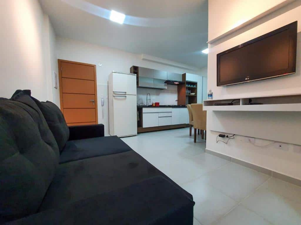 Sala de estar do Apartamento novo. Um sofá do lado esquerdo, no fundo a porta do apartamento. Do lado direito um painel com televisão, no fundo a cozinha com geladeira e a mesa com cadeiras.