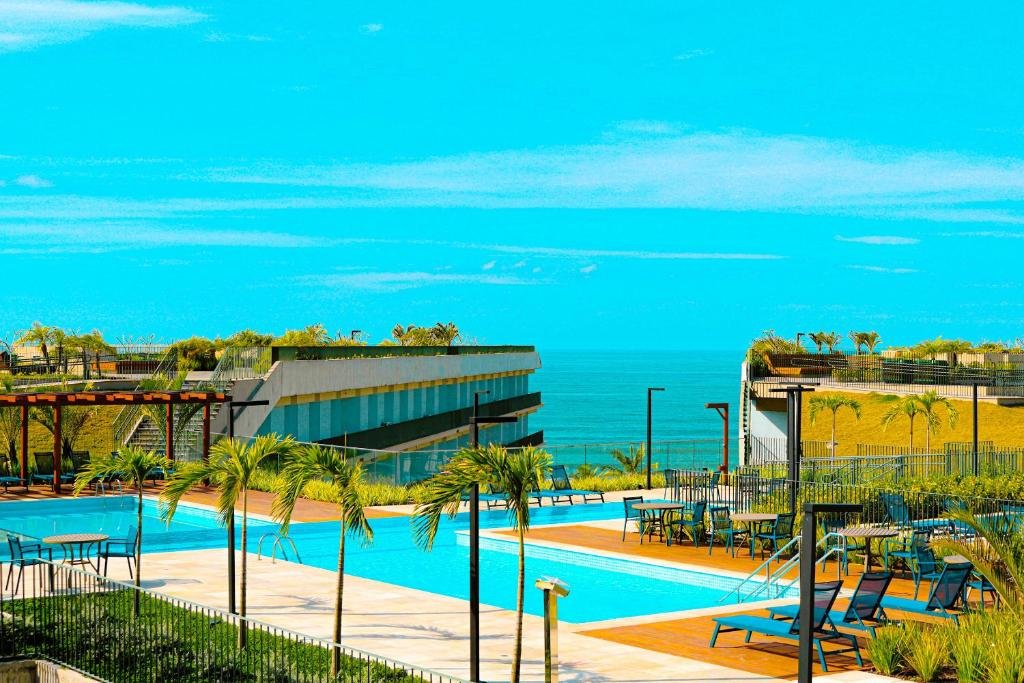 Área de lazer do Apartamento Resort. Uma piscina no terraço, com cadeiras ao redor e palmeiras, do lado direito e esquerdo, no fundo, o condomínio, e a vista da praia no meio.