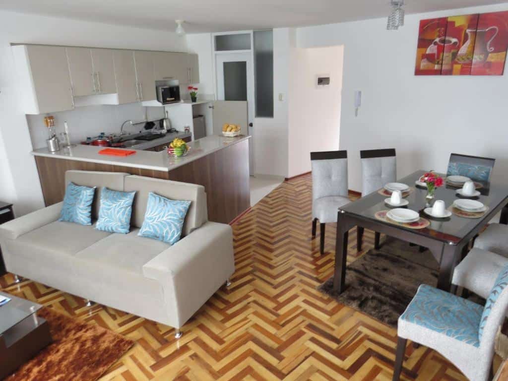 Apartamentos Quewe mostrando a sala de jantar e cozinha aberta. No lado direito há uma mesa de madeira com seis cadeiras, e no lado esquerdo há um sofá de dois lugares, além de um balcão em L e utensilios de cozinha