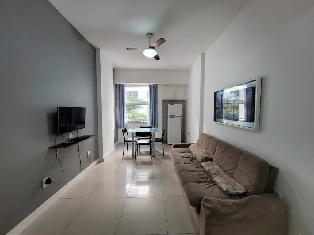 Imagem de uma sala de estar nos Apartments Almirante Gonçalves. A sala de de estar mostra um sofá na direita, e na esquerda, de frente para ele, há uma TV fixa na parede. No fundo há uma mesa com quatro cadeiras , uma janela e uma geladeira com um armário em cima.