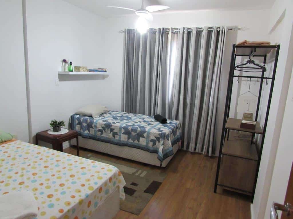 Quarto do Apê da Gabi. Uma cama de casal, um banco e uma cama de solteiro do lado esquerdo, com uma prateleira em cima. Do lado direito uma estante.