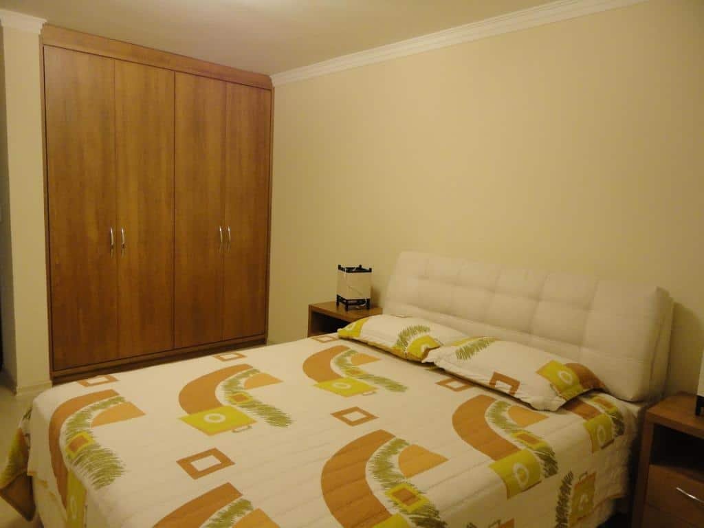 Quarto no Apartamento Baviera. Há uma cama de casal no centro, e uma mesa de cabeceira nos seus dois lados com abajur em cima. Na esquerda há um guarda-roupa. Representa o post sobre airbnb em Canela.