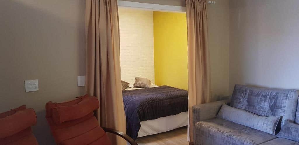 Foto mostrando o quarto no Apto Bell Canela "Cobertura no Centro". Uma cortina separa o quarto, com cama de casal, da sala. Vemos um sofá à direita e outras duas poltronas à esquerda. Entre eles está a área do quarto. Representa o post sobre airbnb em Canela.