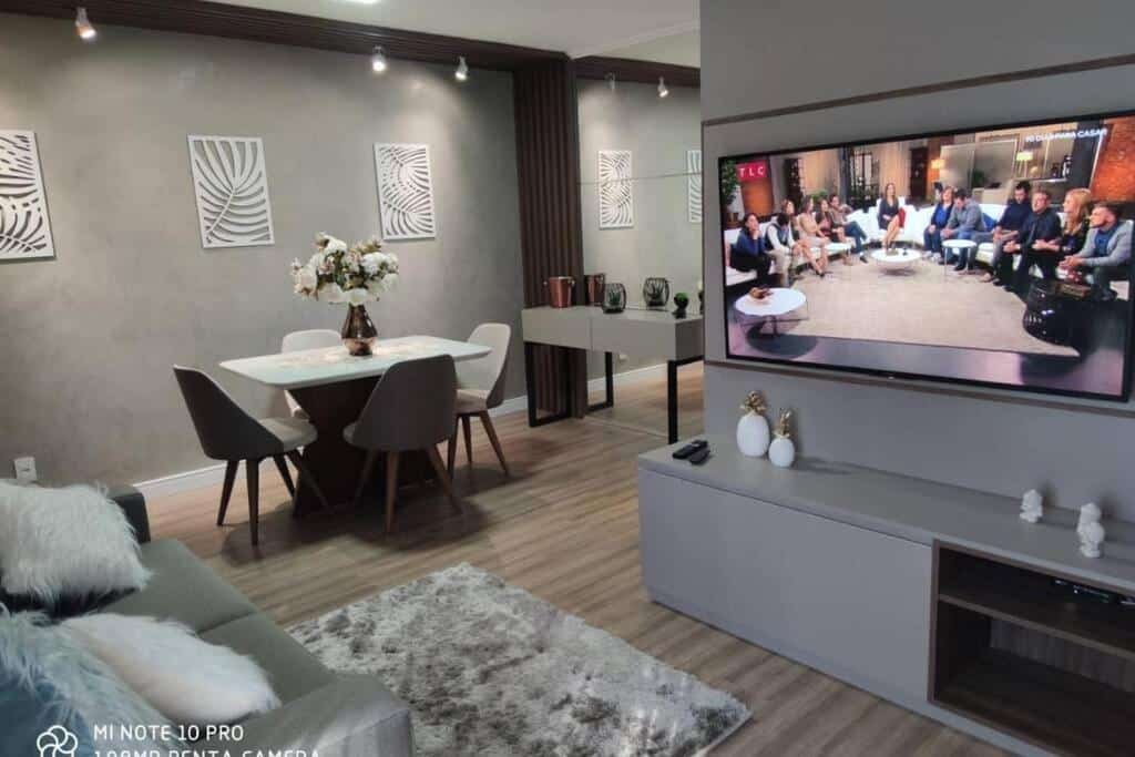 Sala e área de refeições no Lindo apartamento no centro de Canela. O sofá está no canto inferior esquerdo, e na sua frente há um painel com televisão. No fundo, à esquerda,, há uma mesa de jantar com quatro cadeiras.