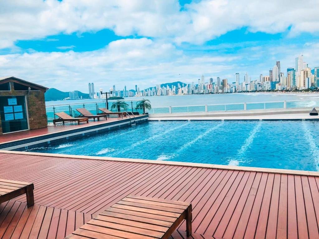 Vista da piscina do Apto 1 dorm frente mar. Cadeiras de tomar sol ao redor, a piscina no meio e no fundo a vista do mar e dos prédios.