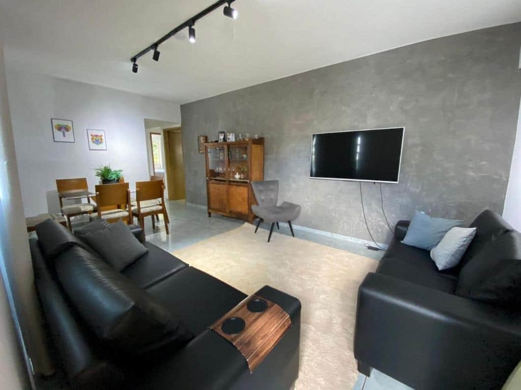 Foto em Apartamento VIP Centro Serra Negra. É uma sala espaçosa com um sofá grande preto no canto inferior esquerdo, e outro igual na direita. De frente para eles há uma parede cinza com uma TV fixa. Debaixo da TV há uma poltrona, e ao lado da poltrona há uma estante. No fundo do ambiente, mais à esquerda, há uma mesa com cadeiras..
