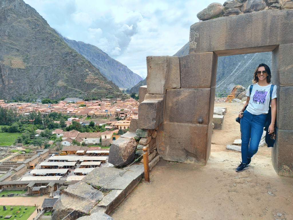 vista panorâmica da cidade de ollantaytambo no vale sagrado dos incas. No lado direito da imagem há um arco de pedra em formato de porta, onde uma mulher está apoiada com os ombros, posando e sorrindo para a câmera. No lado direito da imagem é possível enxergar a cidade lá embiaxo