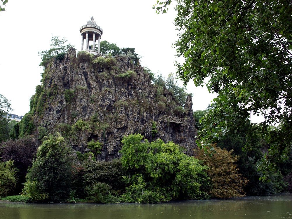 Uma colina rochosa com uma pequena estrutura de templo no topo, o Templo da Sibila, cercada por vegetação exuberante, ao lado de um corpo d