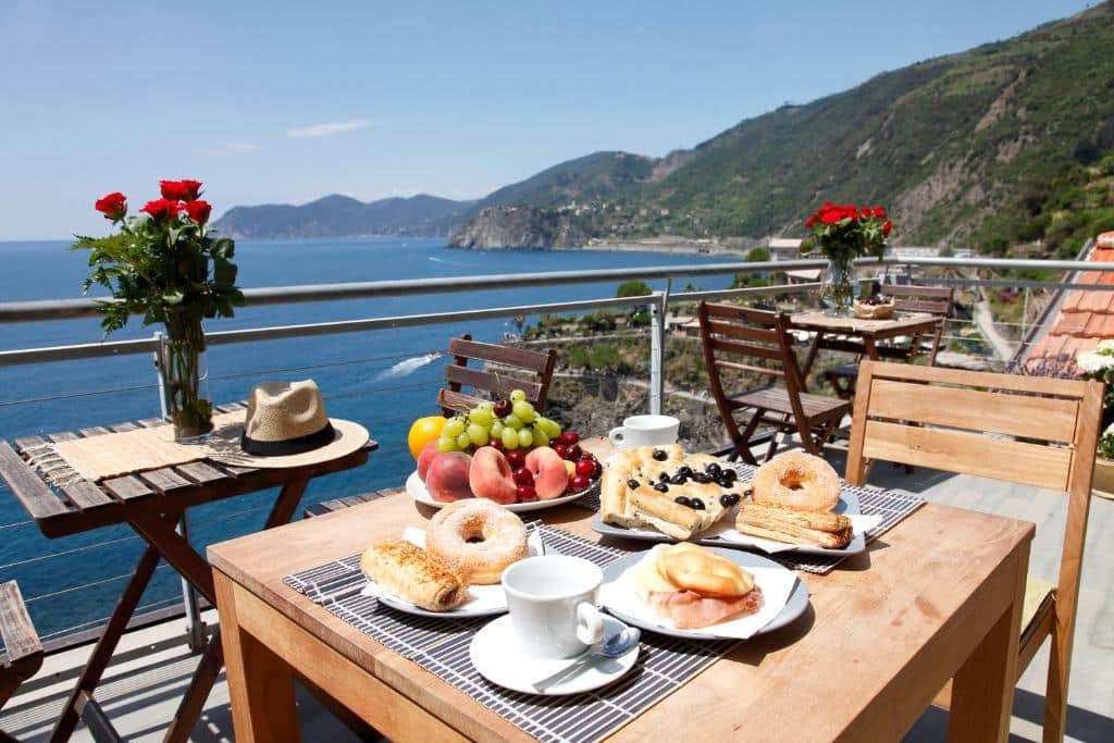 Mesa com cadeira de madeira e algumas comidas em cima, ao fundo outras mesas com flores vermelhas e vista para o mar e a montanha, ilustrando post Hotéis em Cinque Terre.