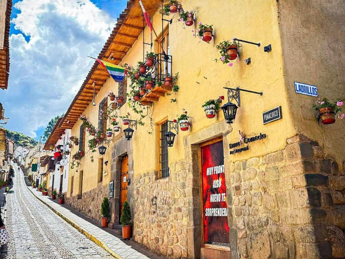 Rua de pedra com um prédio amarelo adornado com plantas em vasos e bandeiras. Uma porta vermelha tem uma frase em espanhol. Céu azul acima.