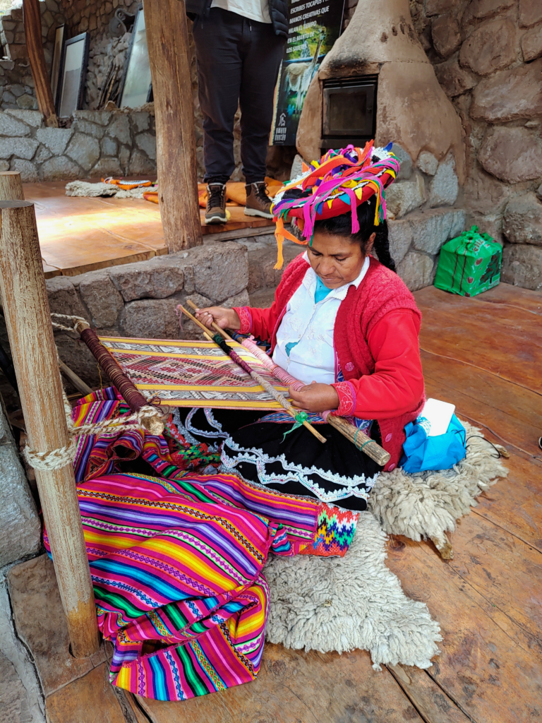 artesã tecendo os fios de lã da mesma forma que seus ancestrais incas costumavam fazer anos atrás. Ela também usa uma roupa típica, com um chapéu vermelho e tiras coloridas presas a ele, e uma manta colorida disposta sobre suas pernas