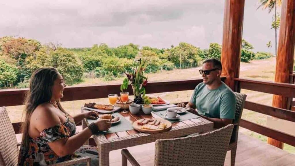 Duas pessoas sentadas em uma mesa ao ar livre desfrutando de um café da manhã com vários pratos e bebidas, com vista para uma paisagem exuberante no Asenza Beach Resort All Inclusive.