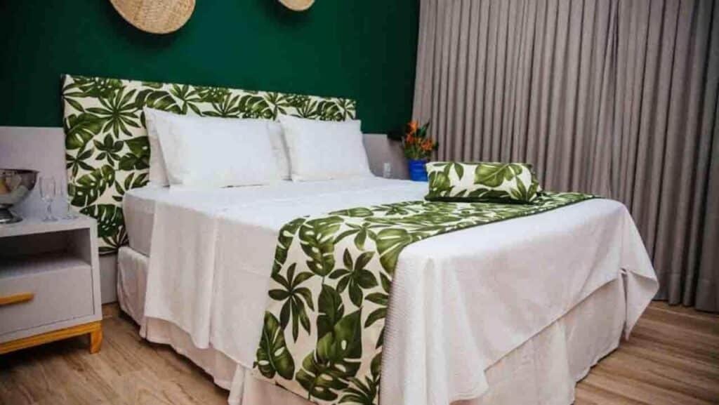 Uma cama bem arrumada com roupa de cama estampada com folhas verdes e brancas, uma mesa de cabeceira com copos e cortinas fechadas ao fundo no Asenza Beach Resort All Inclusive.