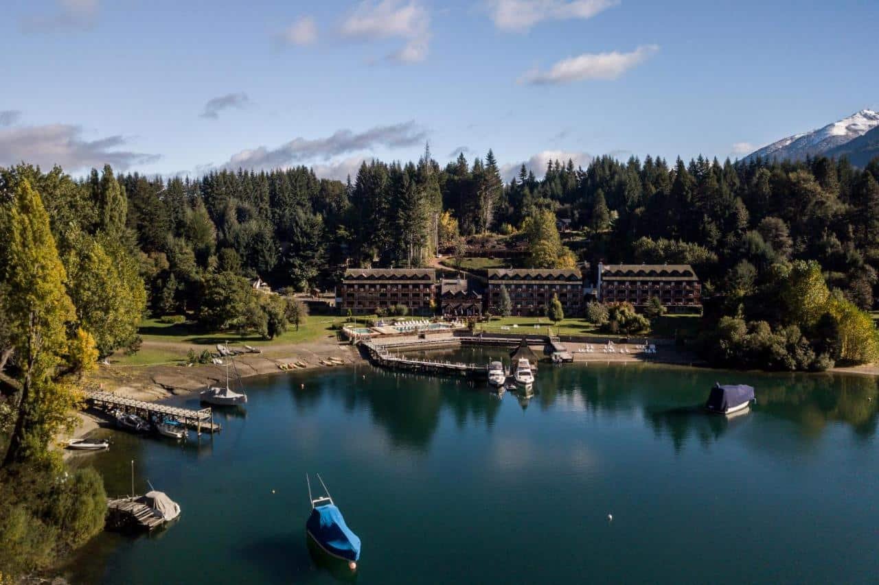 Um tranquilo Bahía Manzano Resort à beira do lago conta com construções de madeira, docas com barcos, cercado por uma densa floresta e uma distante montanha coberta de neve.