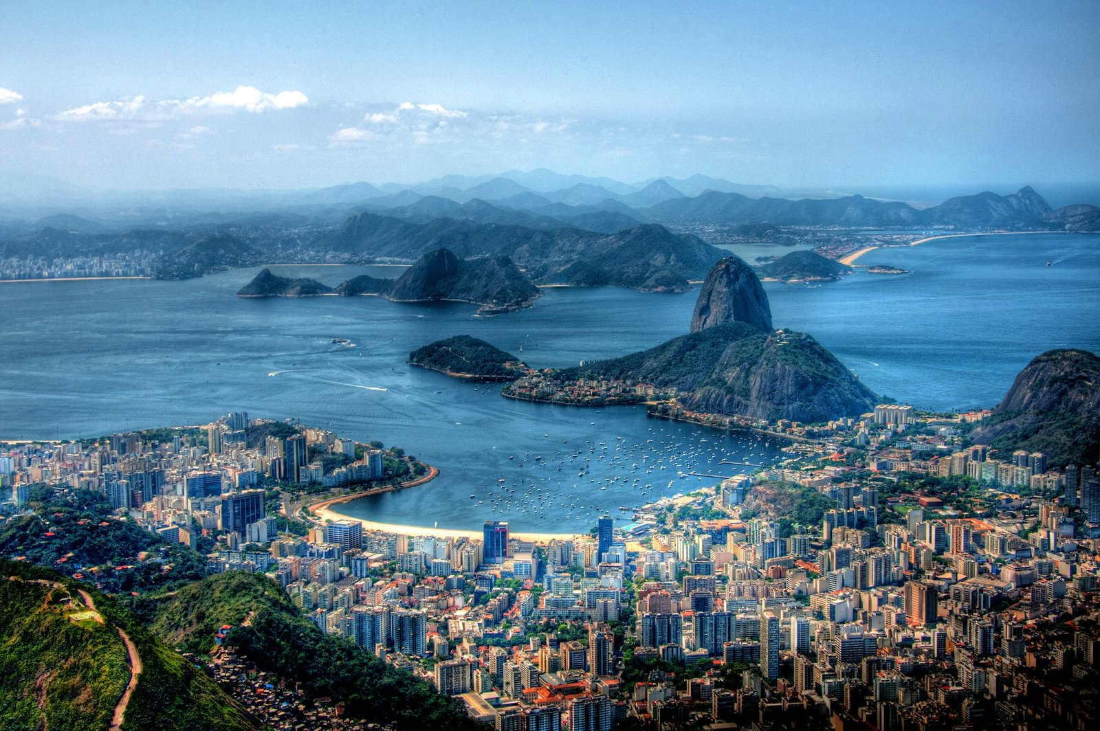 Vista aérea de uma cidade costeira com arranha-céus, praias e montanhas cercadas por águas azuis do oceano sob um céu limpo. Essa é a paisagem de uma das trilhas no Rio de Janeiro.