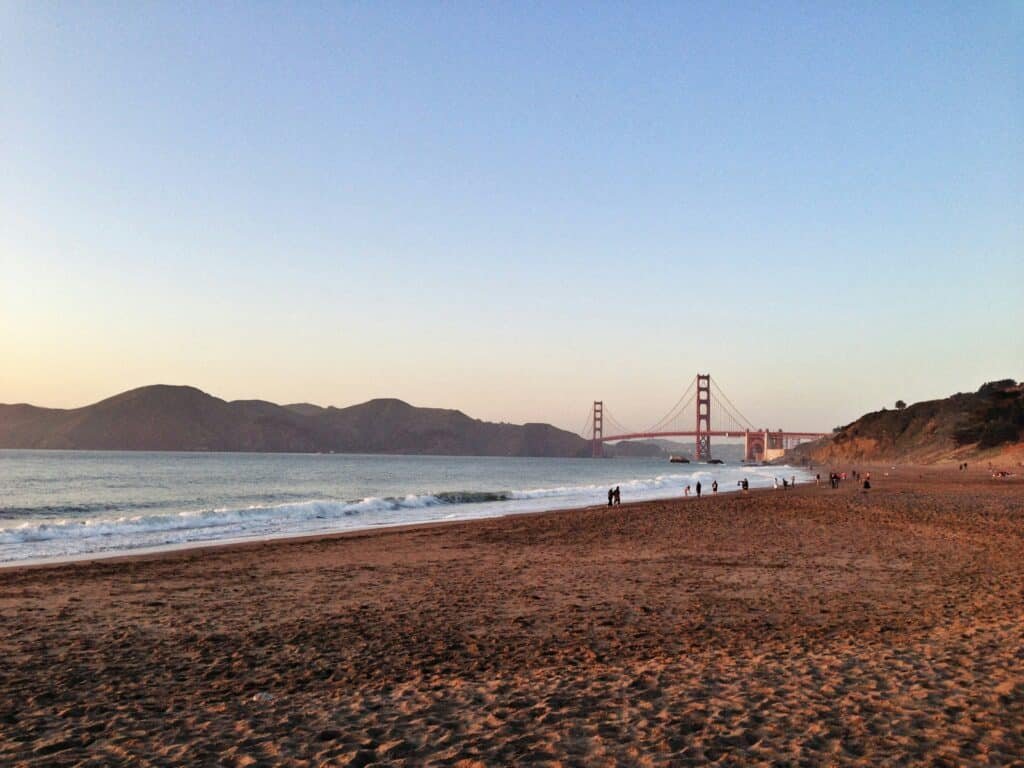 Faixa de areia e mar na praia de Baker Beach. Ao fundo é possível ver a ponte Golden Gate e algumas montanhas.