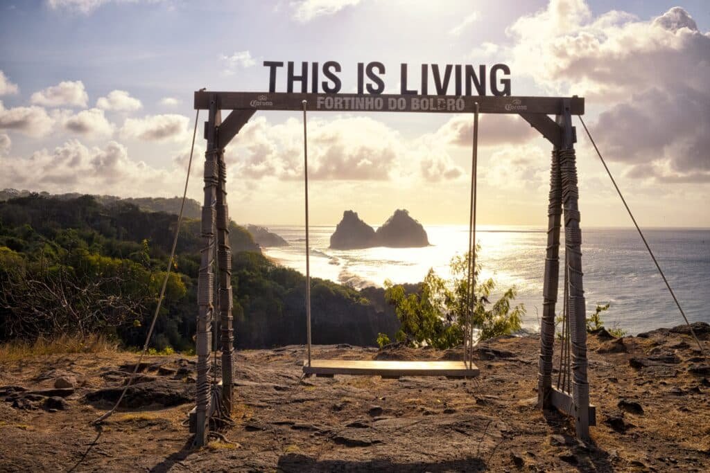 Um balanço de madeira com a placa "ISTO É VIDA" tem vista para o oceano no pôr do sol e duas formações rochosas em um mirante costeiro no Fortinho do Boldró em Fernando de Noronha para representar um dos melhores lugares para lua de mel