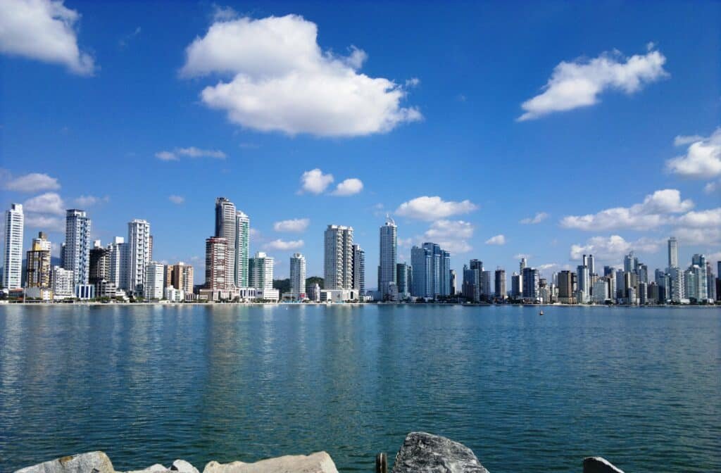 Mar em Balneário Camboriú com prédios de várias alturas na costa. O céu está ensolarado, com poucas nuvens, e há algumas pedras no canto posterior da imagem
