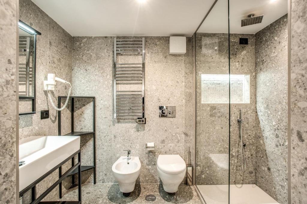banheiro do FOURHEADS Private Suites, com vaso sanitário e bidê, pia com espaço embaixo, prateleira aberta, box averto com chuveirinho, todo revestido em pedra