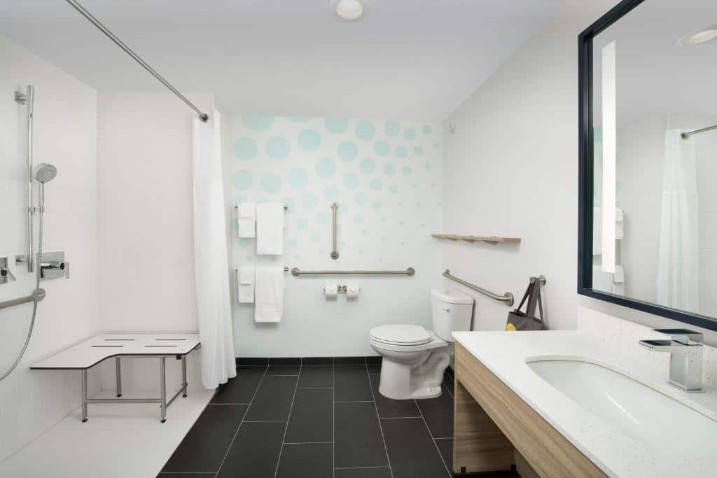 banheiro acessível do Tru By Hilton Orlando Convention Center com ambiente amplo, pia com espaço para cadeiras de rodas com barras de apoio no vaso e no box com cortina, cadeira de banho e chuveirinho