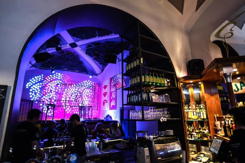 bar do YellowSquare Rome com várias bebidas em uma estante alta e com outros elementos de cozinha, com mesinhas e cadeiras em frente a um palco bastante iluminado em tons de roxo