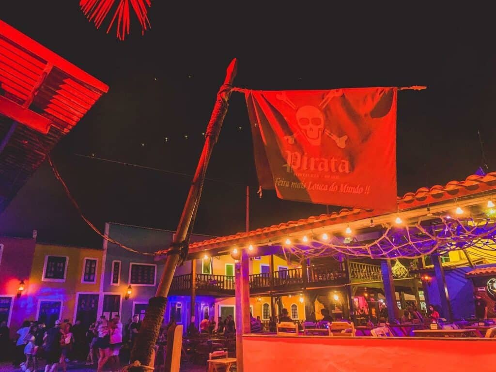 Foto tirada à noite do Bar do Pirata No centro há um mastro com uma bandeira que contém a logo de uma caveira e o nome do bar. trás está o estabelecimento, com luzes vermelhas e violetas. Pessoas estão dançando juntas.