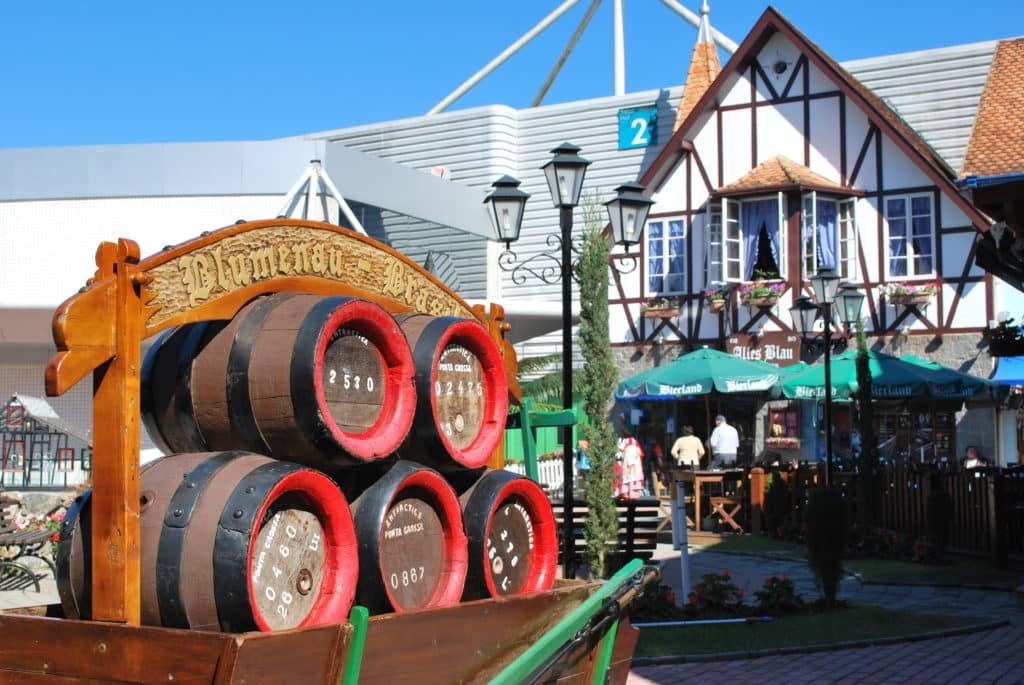 Na imagem vemos monumento decorativo com barris onde há uma placa com os dizeres "Blumenau - Brazil", e ao fundo vemos uma caminha de estilo germânico. Foto do site oficial Parque Vila Germânica.