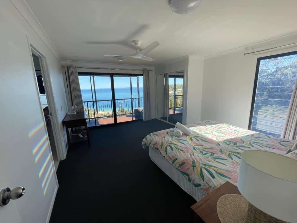 Quarto do Bayview Breeze com uma grande janela com vista para o mar, ventilador de teto, cama com roupa de cama estampada com folhas e uma pequena escrivaninha à esquerda.