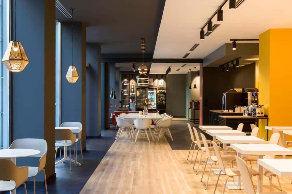 Área de refeições do B&B Hotel Milano Central Station com mesas quadradas e cadeiras, há luminárias pelo local, há uma mesa com cafeteiras e copos, para representar onde ficar em Milão