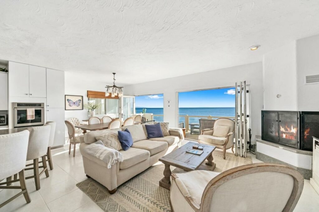 Foto do Beachfront Malibu House para ilustrar post sobre hotéis em Malibu na Cafilórnia. Do lado direito uma sala com lareira, no canto esquerdo a cozinha, no fundo a mesa de jantar. Na parede do fundo duas portas com vista para o mar. Foto para ilustrar post sobre hotéis em Malibu na Califórnia.