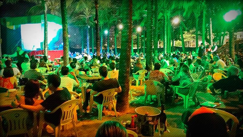 Pessoas sentadas em cadeiras de praia no Beira Mar Grill em Fortaleza. Há coqueiros e um paco na esquerda onde acontece um show.