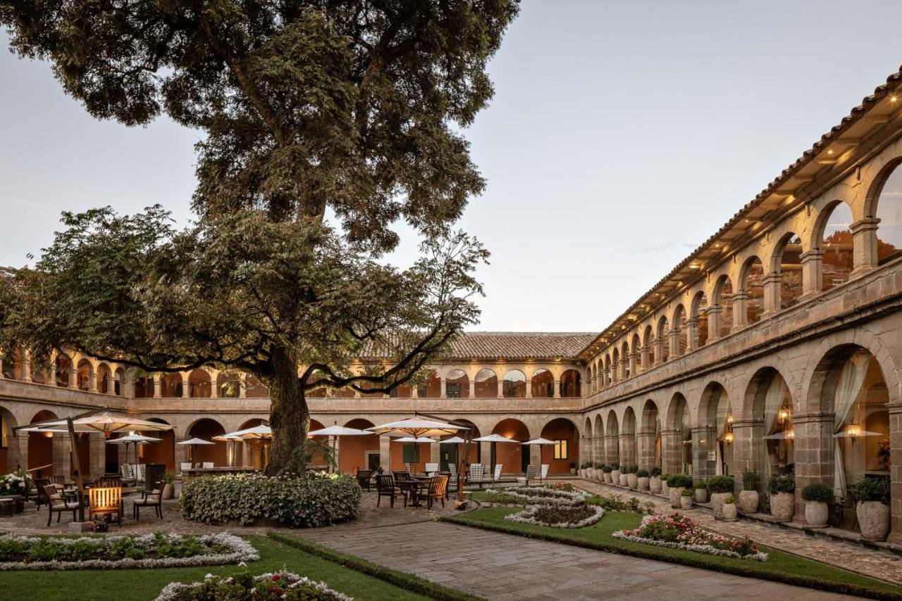 Pátio do Belmond Hotel Monasterio com arcos e colunas, com uma grande árvore, guarda-sóis, mesas, cadeiras e caminhos de jardim bem cuidados. Imagem para ilustrar post sobre réveillon em Cusco.