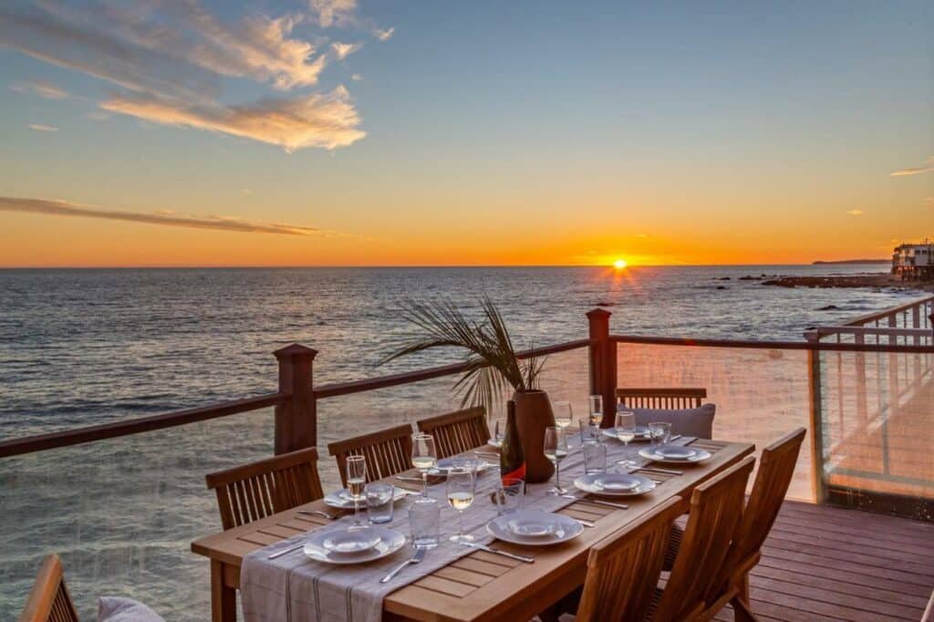 Varanda do Blue Dolphin: An Oceanfront Malibu Sanctuary. Uma mesa posta de jantar, no fundo a vista do pôr do sol com o mar. Foto para ilustrar post sobre hotéis em Malibu na Califórnia.