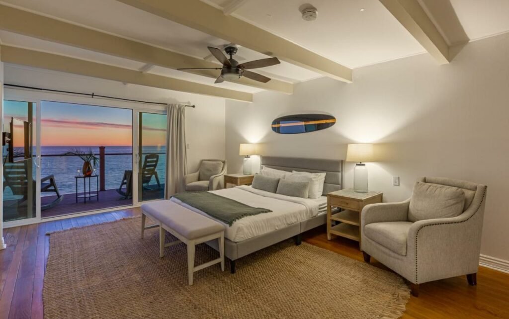 Quarto do Blue Dolphin: An Oceanfront Malibu Sanctuary. Do lado direito uma cama de casal, de cada lado uma cômoda com abajur e uma poltrona. Na frrente um sofá. Do lado esquerdo, no fundo uma varanda com vista do mar e duas cadeiras.