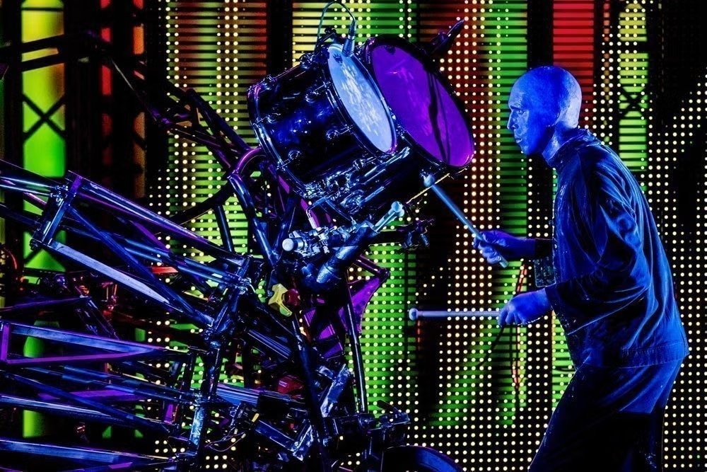 Um dos integrantes do Blue Man Group todo pintado de azul toca uma bateria. O fundo é todo colorido.