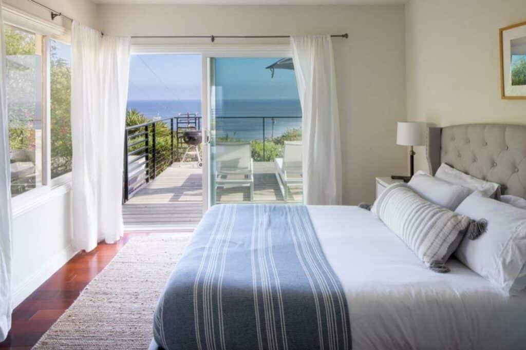 Quarto do Bluff by AvantStay Gorgeous. Do lado direito uma cama de casal, no fundo uma cômoda com um abajur, de frente pra cama uma janela. No fundo a porta da varanda com vista do mar.