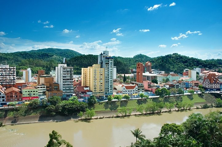 Rio em Blumenau com várias casas e diferentes prédios em ruas costeiras e várias montanhas verdes no horizonte durante um dia ensolarado, com o céu azul e algumas nuvens brancas