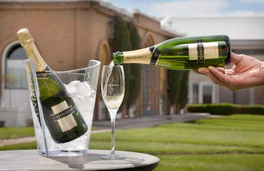Na imagem vemos duas garrafas de espumante Chandon, uma em balde com gelo, e outra sendo servida em taça. Ao fundo está a bodega Chandon. Foto do facebook da bodega. - Vinicola Mendoza