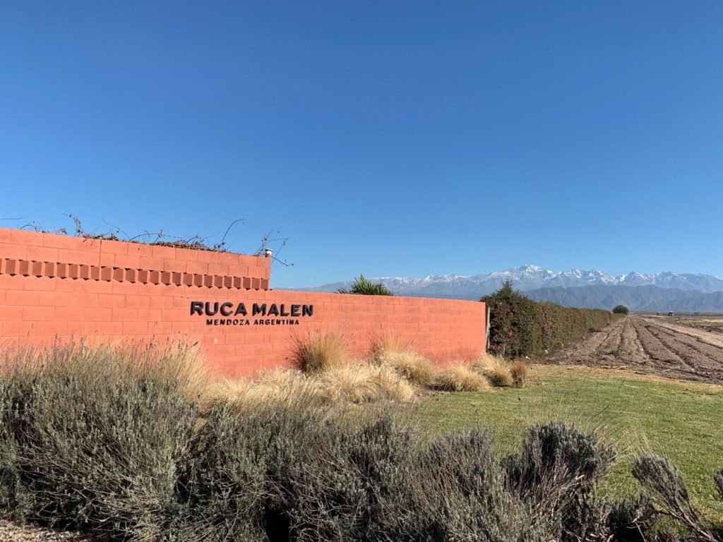 Vista de muro da fachada da bodega Ruca Malen em Mendoza, com plantações vitivinícolas e montanhas ao fundo, em dia de céu azul limpo. Foto de Bruno Tavares.